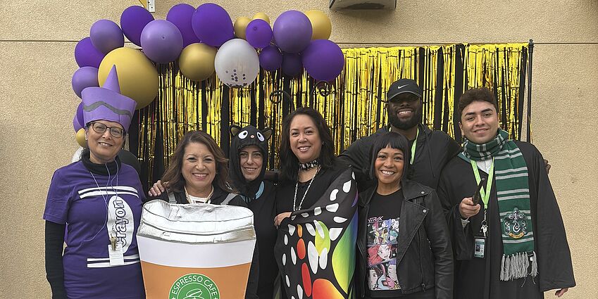 Adults in various Halloween costumes in front of colorful balloons.