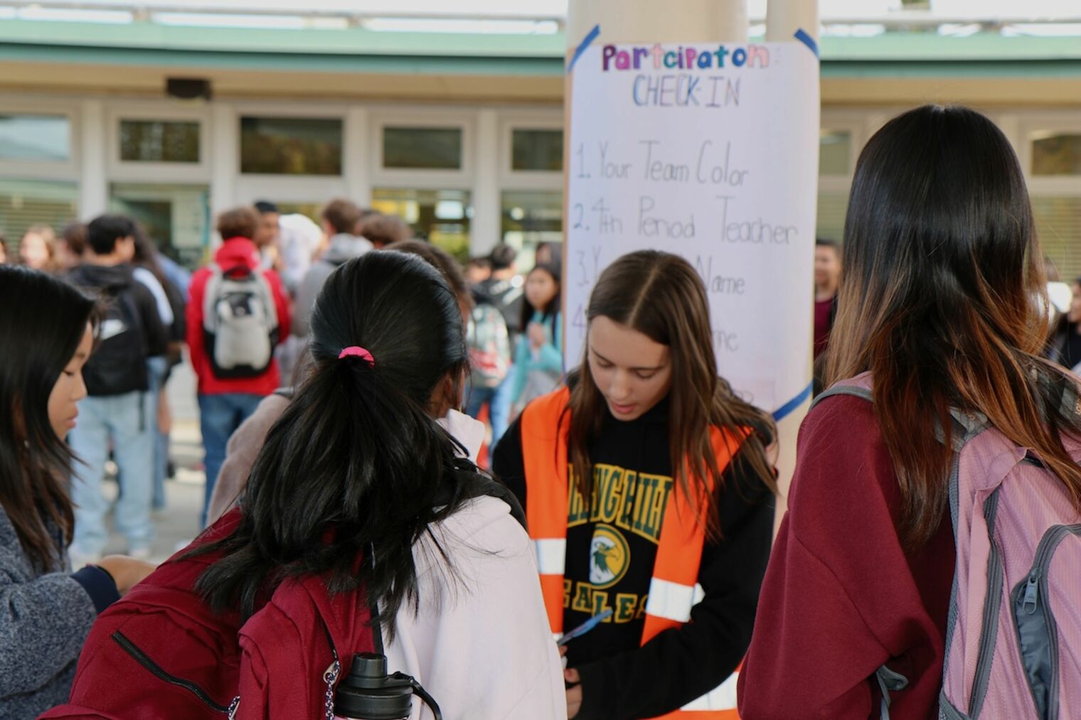 middle school girl wearing an orange safety vest stands at a participation sign up station as several students stand waiting to sign up