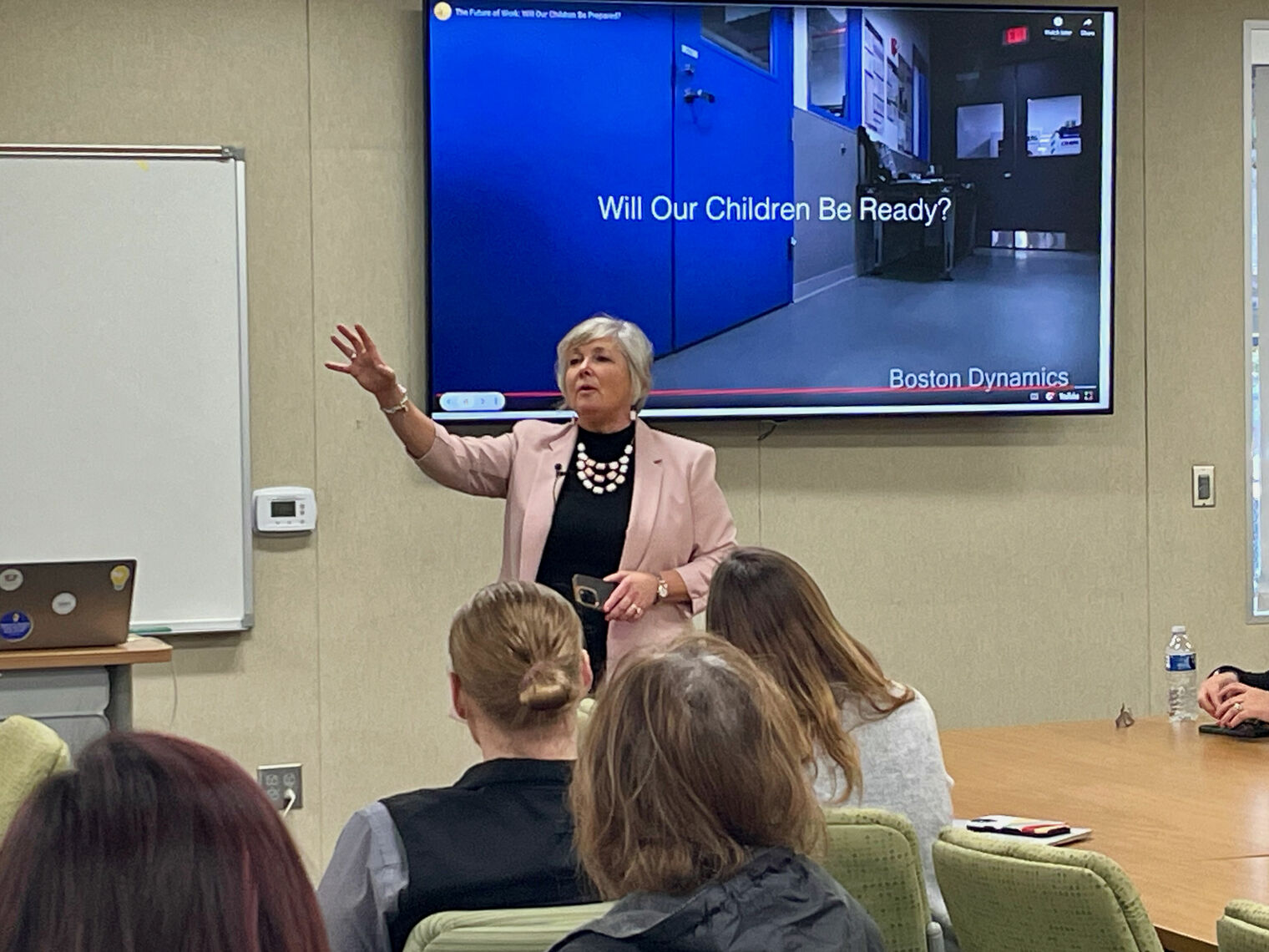 woman with raised hand speaks to a group of adults. behind her on screen it reads 