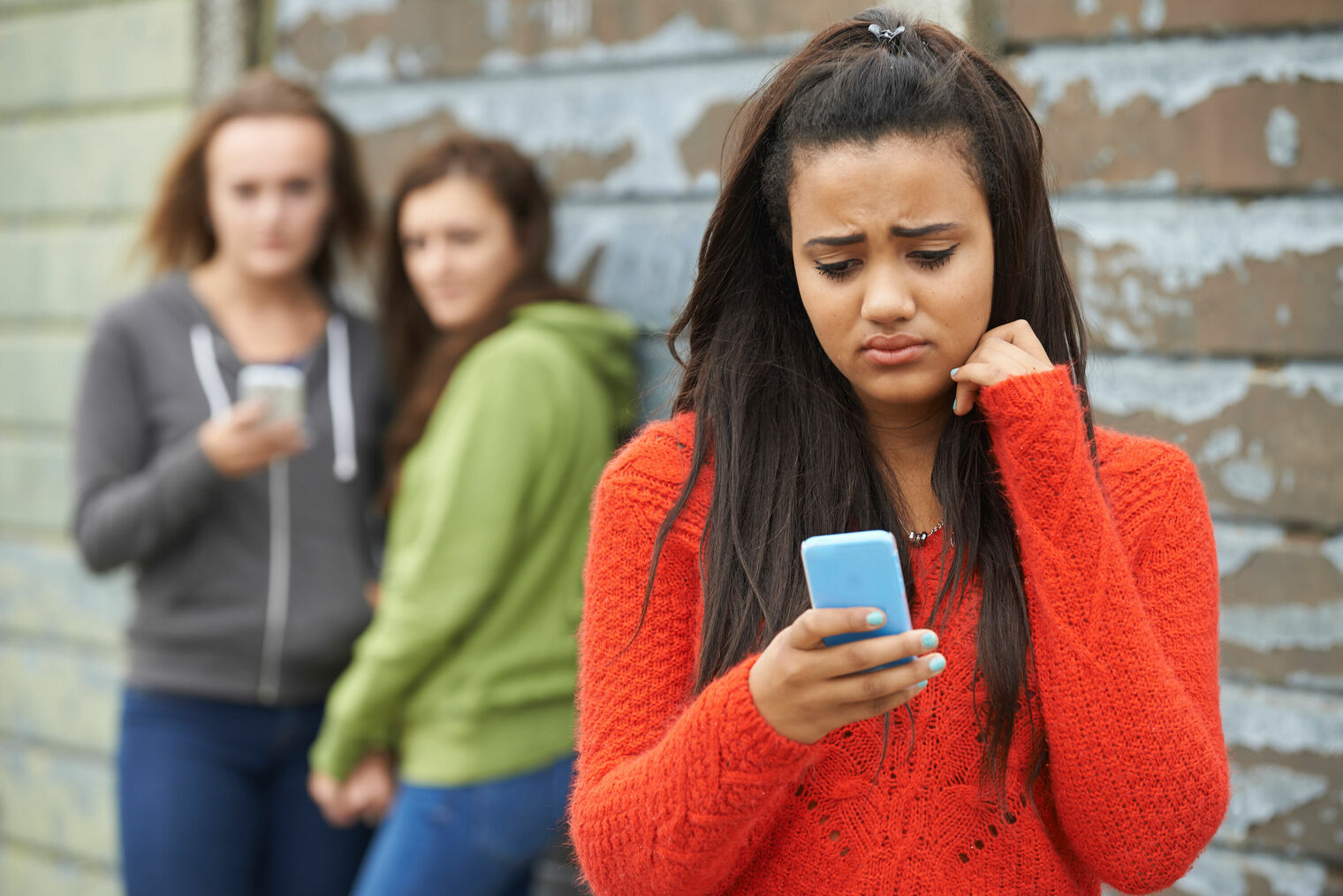 sad teen in foreground looking at cell phone as 2 girls in background look on with sneers.